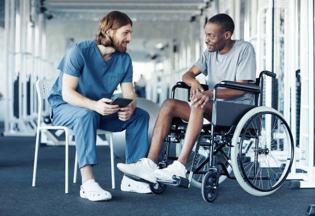 A person in medical scrubs sitting on a chair talking to another person in a wheelchair